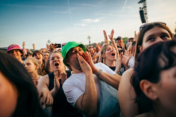 Crowd of people at an outdoor event cheering and clapping during daytime with a man in a green hat shouting.