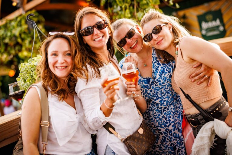 Four smiling women wearing sunglasses and casual clothes, holding drinks, posing closely together outdoors.