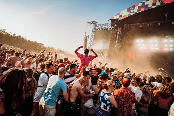Crowded outdoor music festival with people dancing and cheering near a stage during sunset.