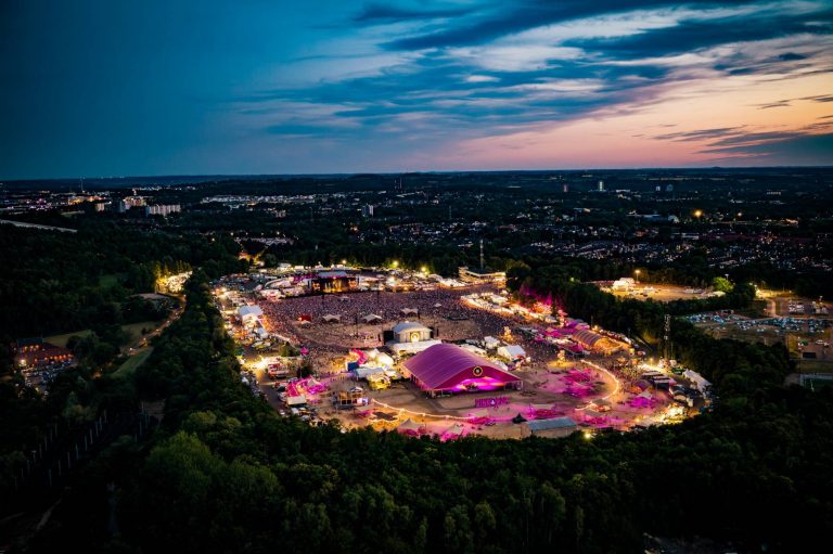 Aerial view of a large outdoor festival at dusk with bright lights, a crowd, and tents surrounded by trees and cityscape.