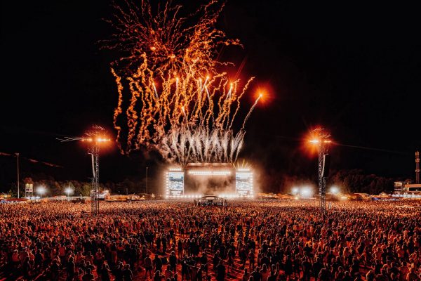Large crowd at a nighttime outdoor festival watching fireworks above a brightly lit main stage.