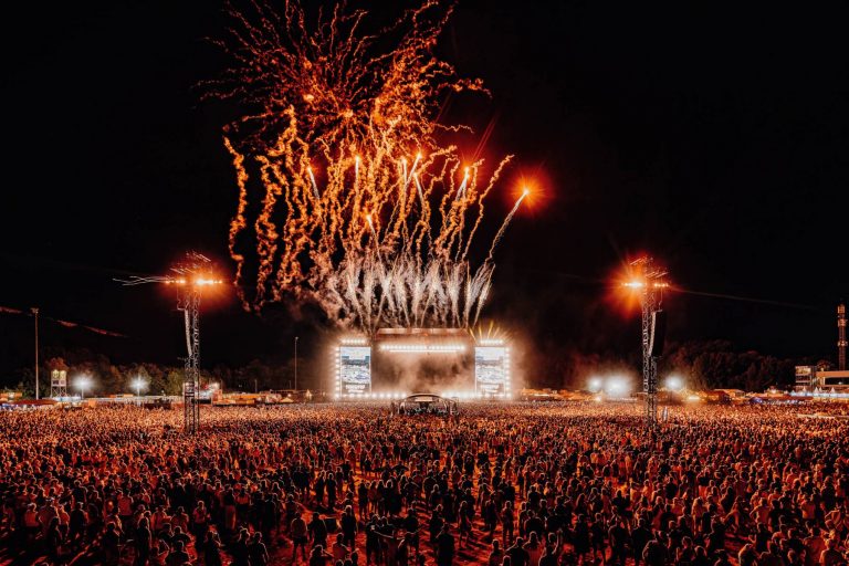 Large crowd at a nighttime outdoor festival watching fireworks above a brightly lit main stage.