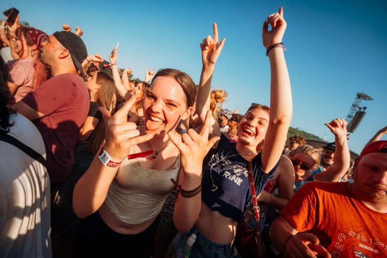 Young woman at a crowded outdoor festival making hand signs and smiling with tongue out, surrounded by other joyful attendees.