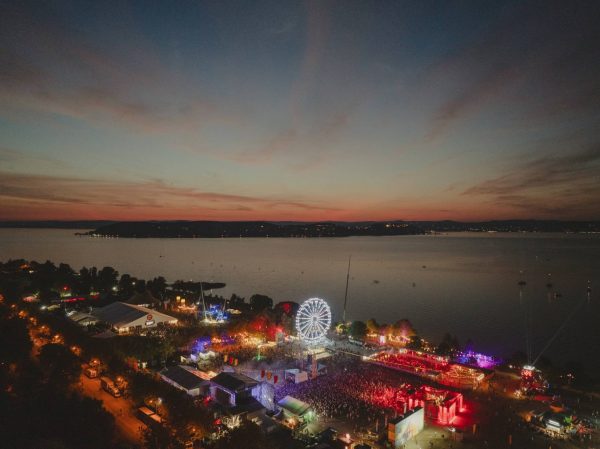 Aerial view of a large outdoor festival at dusk by a lake, featuring a brightly lit Ferris wheel and colorful crowd lights.