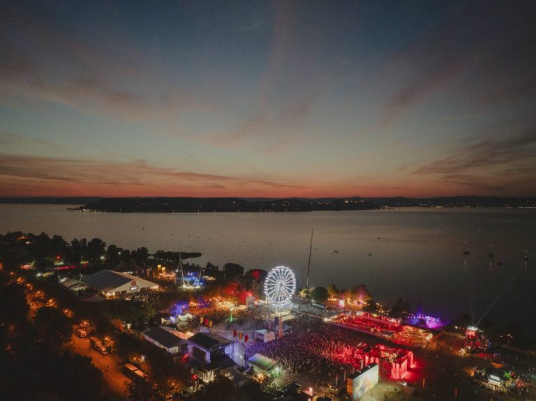 Aerial view of a large outdoor festival at dusk by a lake, featuring a brightly lit Ferris wheel and colorful crowd lights.