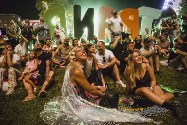 Diverse crowd sitting on grass at night looking up, with large colorful letters spelling 