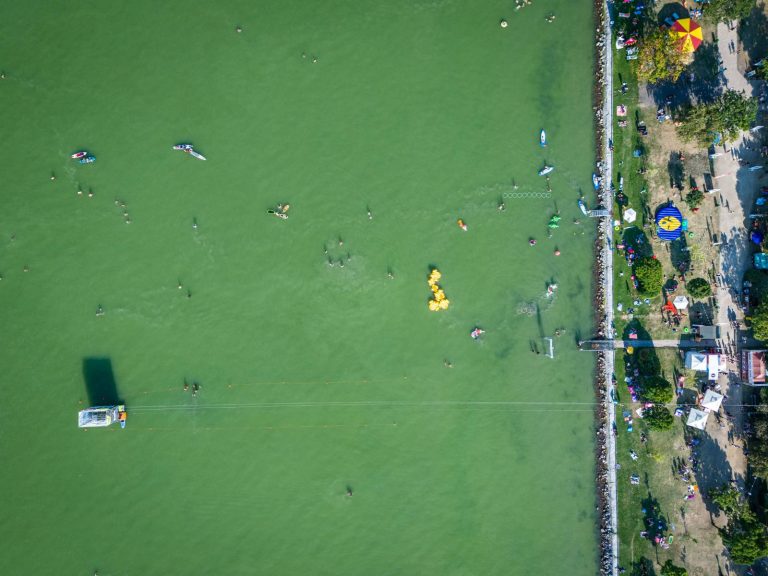 Aerial view of people swimming and using inflatable water toys in a lake near a grassy shore with umbrellas and trees.