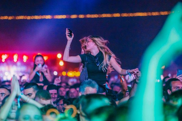 Young woman with long blonde hair dancing and holding a phone above a crowd at a nighttime music festival or concert.