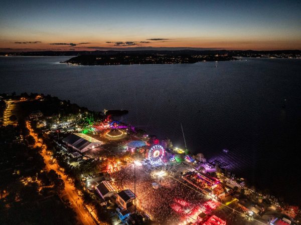 Aerial view of a large outdoor festival near a body of water at dusk, with a brightly lit Ferris wheel and crowds of people.