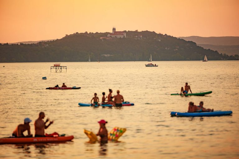 People kayaking and swimming on a lake at sunset with a hill and building in the background