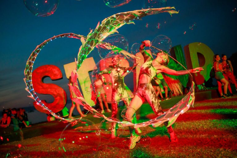Two performers in tie-dye costumes creating large glowing bubble rings with a crowd and colorful letters in the background at dusk.