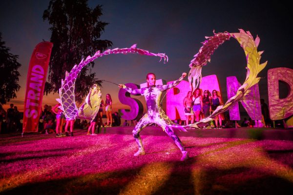 Performer in patterned bodysuit waving large dragon-shaped ribbons at outdoor festival with crowd and 