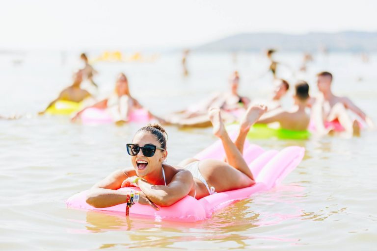 Woman in sunglasses smiling and lying on a pink inflatable float in the water, with others floating in the background.