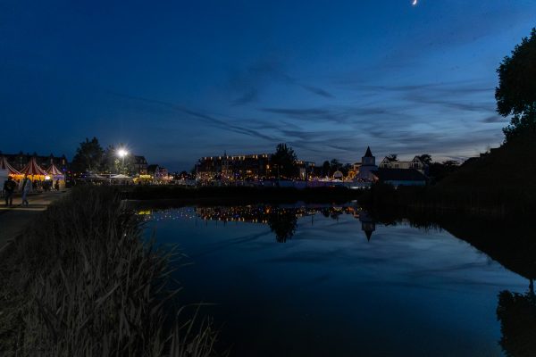 Night scene of a carnival or fair near a calm body of water, with colorful lights reflecting on the surface and a crescent moon in the sky.