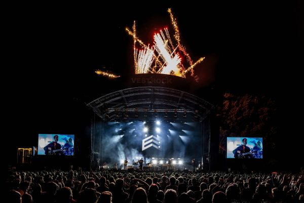 Outdoor night concert with a large crowd, band performing on stage under bright lights and fireworks overhead.