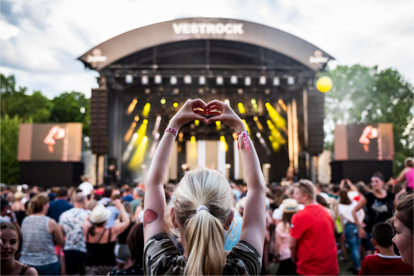 Person with blonde hair making a heart shape with hands at a crowded outdoor music festival stage with 
