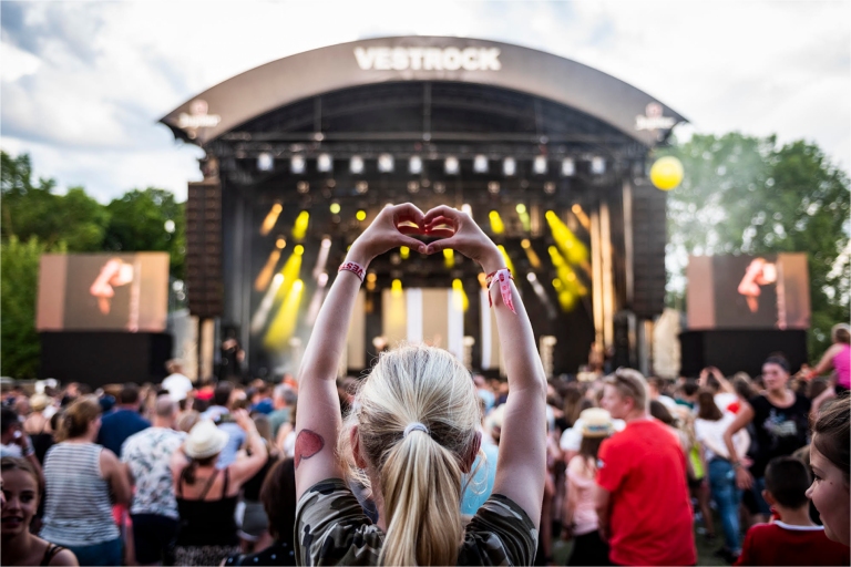 Person with blonde hair making a heart shape with hands at a crowded outdoor music festival stage with 