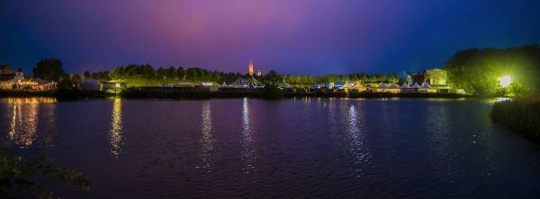 Nighttime view of a lakeside festival with illuminated tents, a large striped circus tent, trees, and a church tower in the background.
