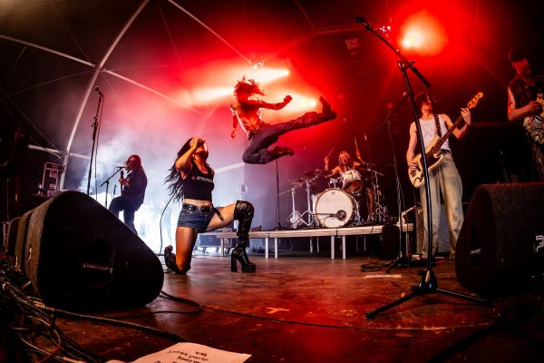 Rock band performing on stage with energetic singer kneeling and shirtless man jumping mid-air under red lights.