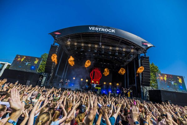 Crowd with raised hands watching live music performance on Vestrock festival stage with pyrotechnics under clear blue sky