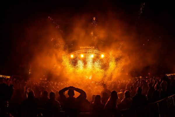 Crowd watching a nighttime concert with bright orange stage lights and rising sparks at the Vestrock festival.