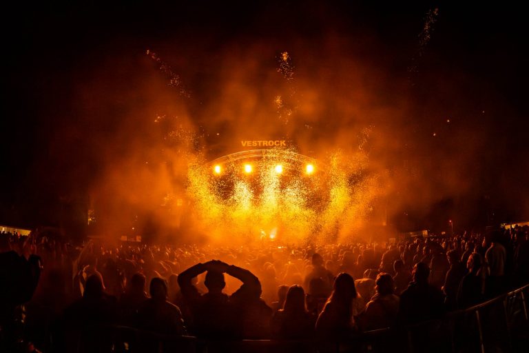 Crowd watching a nighttime concert with bright orange stage lights and rising sparks at the Vestrock festival.