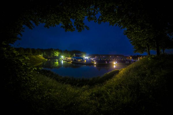 Nighttime view of a lit campsite with tents by a calm lake, framed by dark trees and grass.