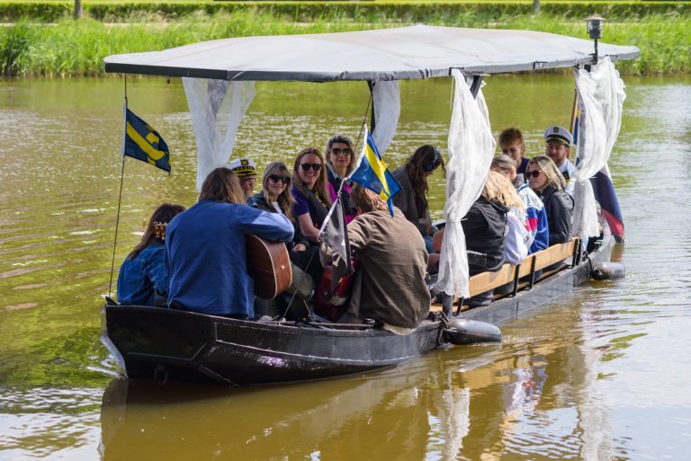 People sitting on a covered boat with lace curtains, some wearing sunglasses and captain hats, on a calm river.