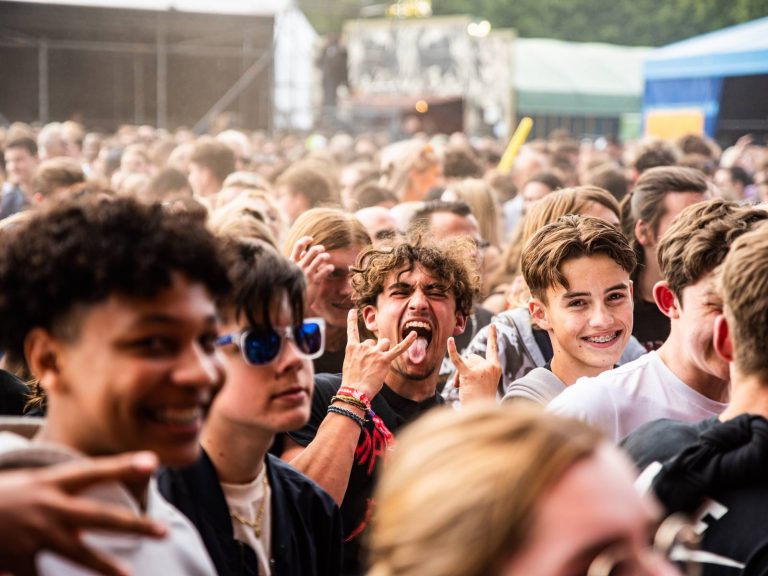 Young men smiling and making hand gestures in a crowded outdoor event setting with tents in the background