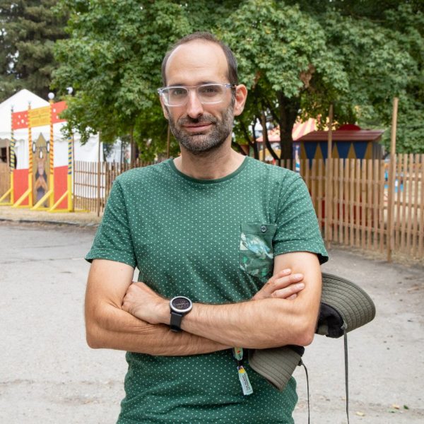 Man with glasses and a short beard wearing a green dotted t-shirt, crossing his arms and holding a wide-brimmed hat outdoors with trees in the background.
