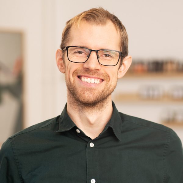Smiling man with light brown hair, glasses, and a short beard wearing a dark green button-up shirt against a blurred indoor background.