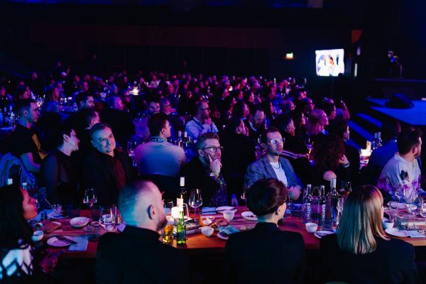 Audience seated at tables with drinks and food, watching a stage event in a dimly lit room with blue and purple lighting