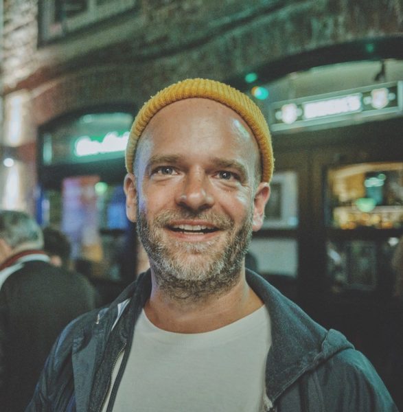 Man with a beard wearing a yellow beanie and jacket, smiling in an indoor bar or pub setting at night.