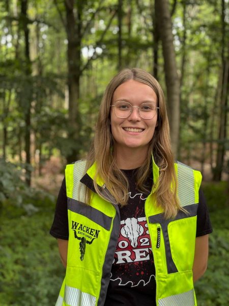 Young woman wearing glasses and a high-visibility vest standing in a forested area, smiling at the camera.