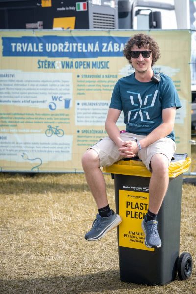 Man wearing sunglasses sitting on a recycling bin labeled for plastic and metal at an outdoor event with a music festival banner in the background.