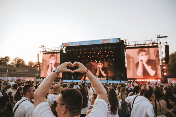 Crowd at outdoor concert with a person in the foreground making a heart shape with their hands facing the stage.