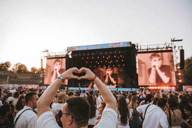 Crowd at outdoor concert with a person in the foreground making a heart shape with their hands facing the stage.