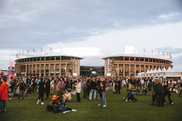 Large crowd gathered on grass outside a stadium with flags, tents, and signs during an outdoor event.