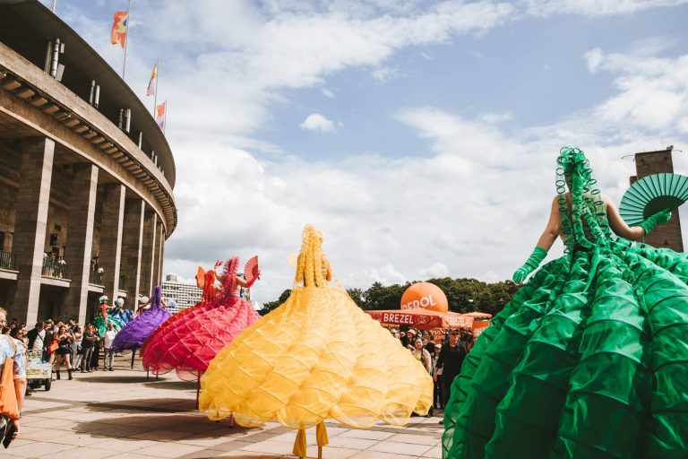 Performers in colorful, voluminous dresses with fans stand in front of a large curved building during an outdoor event.