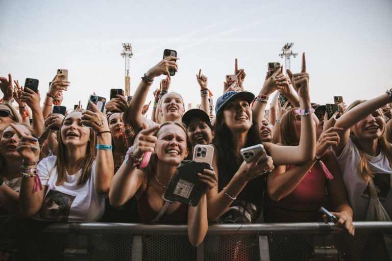 Excited crowd of young people holding smartphones and cheering at an outdoor event or concert.