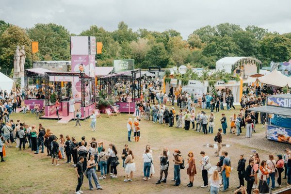 Large crowd forming lines at an outdoor event with booths and greenery in the background under a cloudy sky