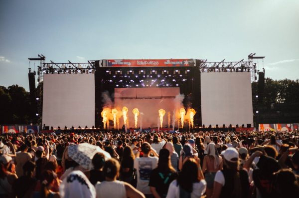 Large outdoor music festival crowd watching a stage with multiple flames shooting up and bright lights overhead under a clear sky.