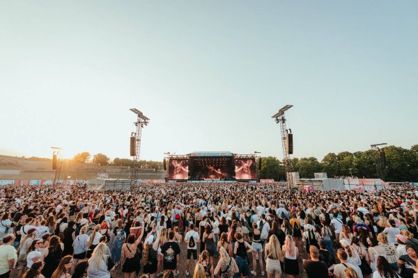 Large outdoor music festival crowd watching a performance on a distant stage with two large screens at sunset