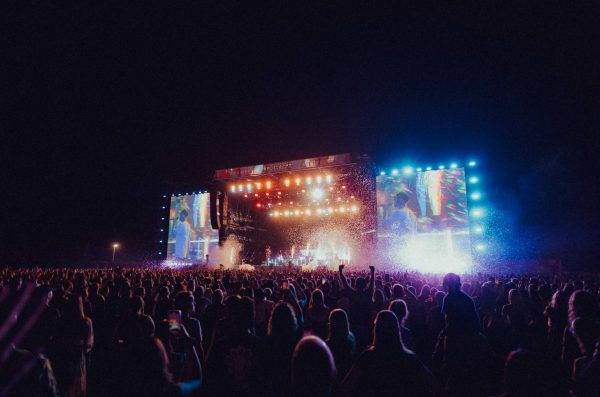 Large crowd at an outdoor night concert with bright stage lights and large screens showing performers.