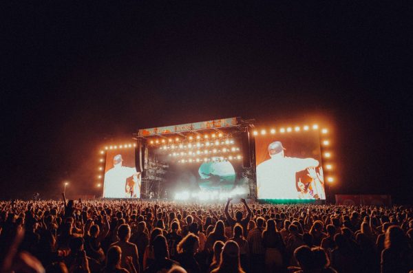 Large crowd at a nighttime outdoor concert with a brightly lit stage and two large screens showing a performer in a white shirt and cap.