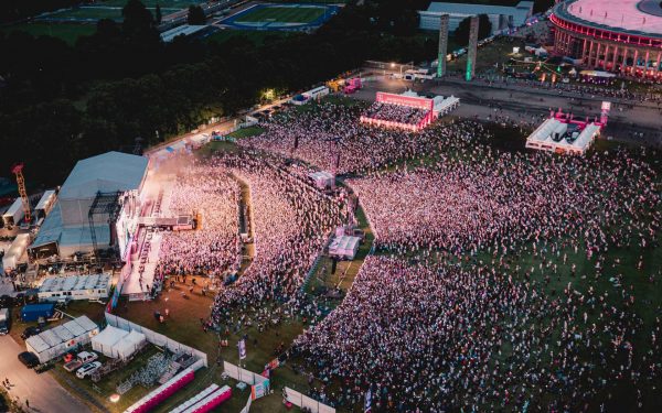 Aerial view of a large outdoor concert with thousands of people gathered in front of a brightly lit stage at dusk.