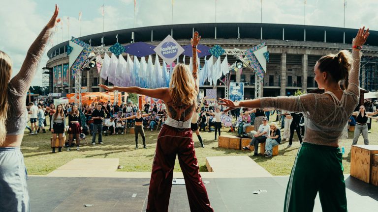 Three women leading an outdoor dance or fitness session on a stage in front of a crowd at a Festival Palooza event.