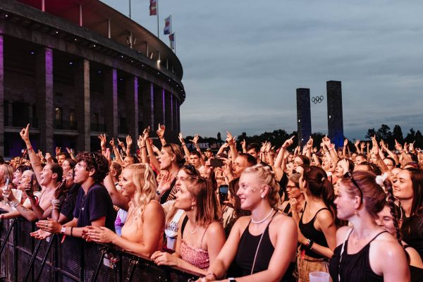 Excited crowd of young people enjoying an outdoor concert near a large stadium with flags and Olympic rings in the background at dusk