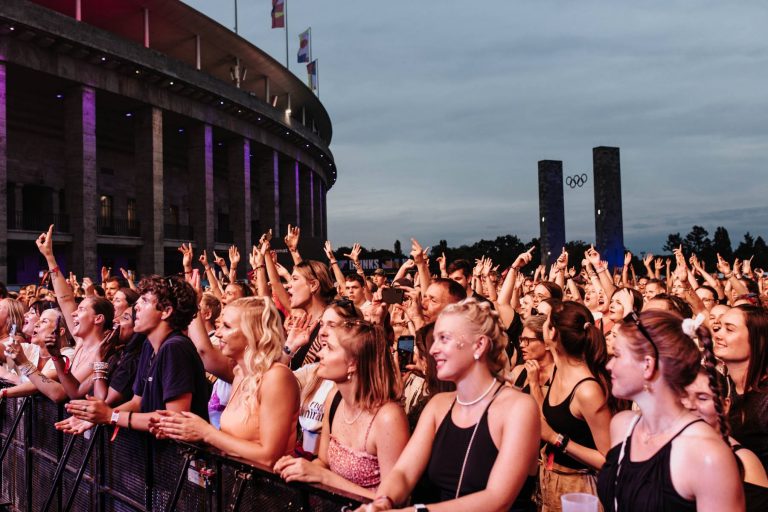 Excited crowd of young people enjoying an outdoor concert near a large stadium with flags and Olympic rings in the background at dusk
