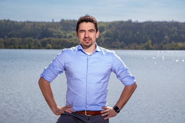 Man in a light blue shirt with rolled-up sleeves standing with hands on hips in front of a lake and forest background.